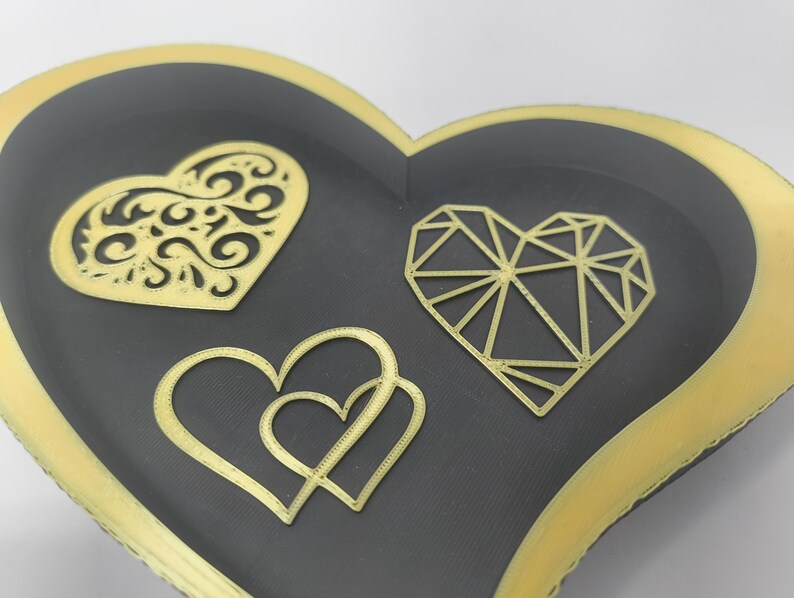 May include: A heart-shaped decorative tray with a black interior and gold trim. The tray features three gold heart designs: a filigree heart, a geometric heart, and two intertwined hearts. The tray is likely used for jewelry or small items.