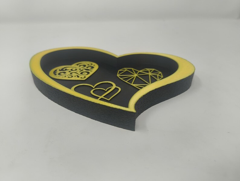 May include: A heart-shaped decorative tray with a black interior and a yellow border. The tray features three heart designs: a filigree heart, a geometric heart, and two interlocking hearts.