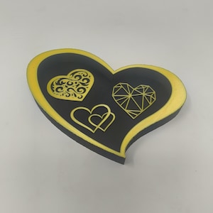 May include: A heart-shaped decorative tray with a black interior and a gold-colored border. The tray features three heart designs in gold: a filigree heart, a geometric heart, and two interlocking hearts.