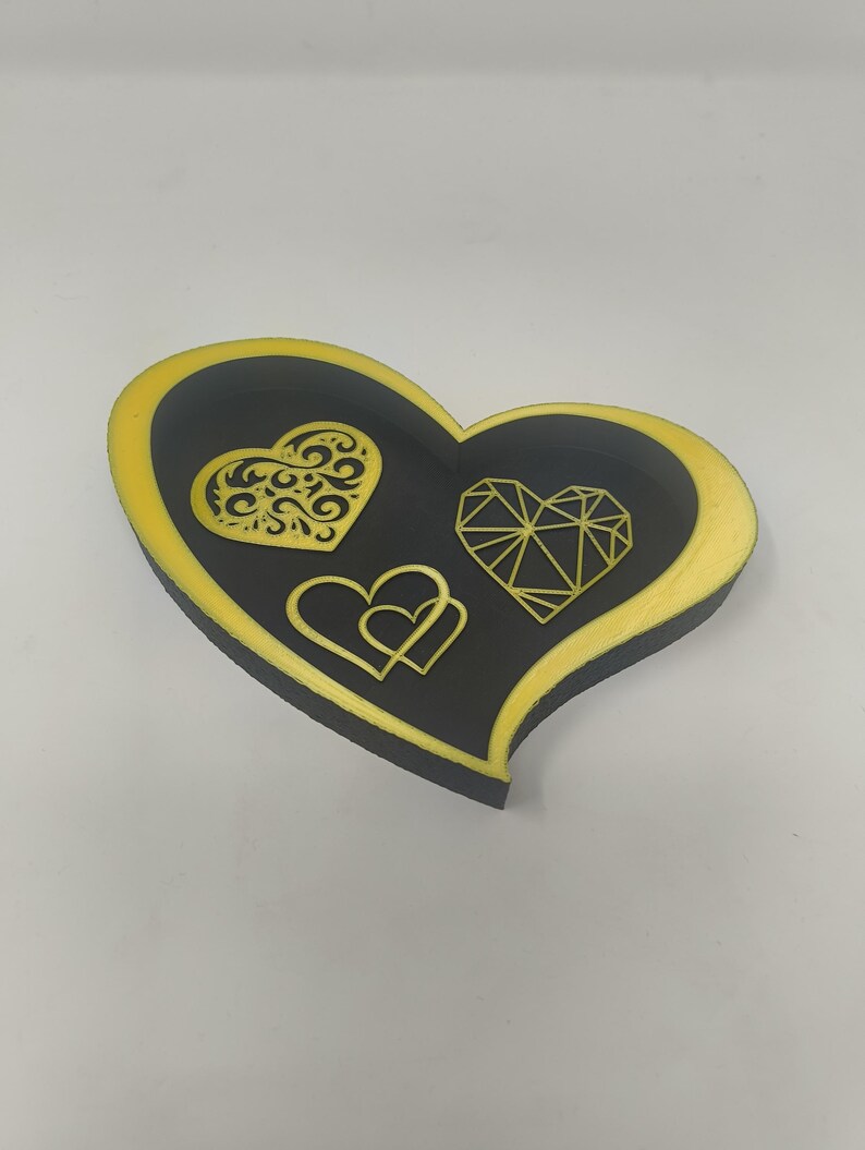 May include: A heart-shaped decorative tray with a black interior and a gold-colored border. The tray features three heart designs in gold: a filigree heart, a geometric heart, and two interlocking hearts.