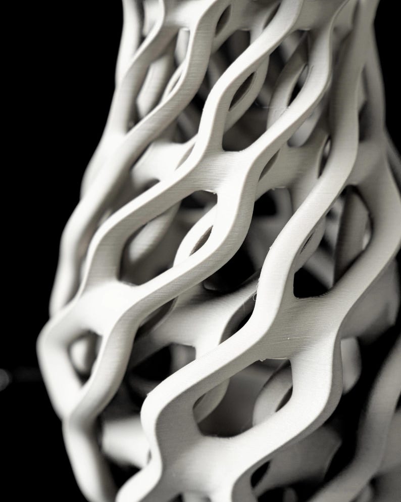 May include: A white, abstract sculpture with a complex, interwoven design. The form appears to be a vase or decorative object, featuring a series of curved, flowing lines that create open spaces. The piece is set against a black background, highlighting its texture and form.