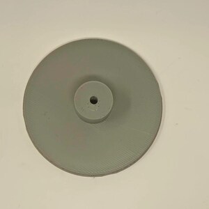 May include: A light gray, circular object with a raised, cylindrical center. The object has a ridged texture on the outer disc and a hole in the center cylinder. The object is likely a component or part of a larger mechanism.