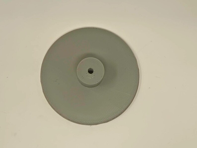 May include: A light gray, circular object with a raised, cylindrical center. The object has a ridged texture on the outer disc and a hole in the center cylinder. The object is likely a component or part of a larger mechanism.
