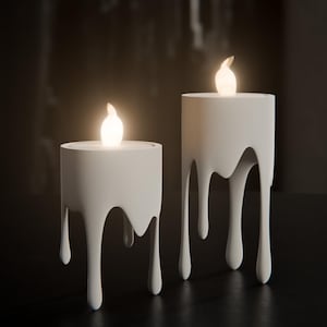May include: Two white candle holders with a melted wax design. Each holder has a glowing, flame-shaped light at the top. The holders are on a dark surface, creating a warm, inviting ambiance. Home decor.