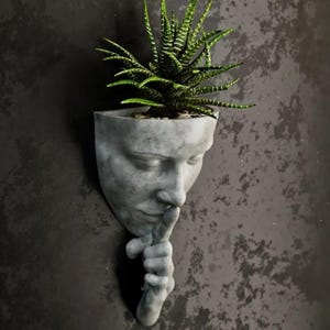 May include: A grey concrete planter shaped like a human face with its finger to its lips, as if to say 'shhh'. A green succulent plant is growing out of the top of the planter.