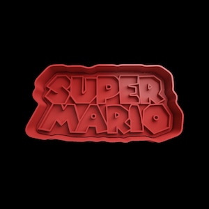 Puede incluir: Cortador de galletas de pl&aacute;stico rojo con forma del logotipo de Super Mario. El logotipo est&aacute; en una fuente en negrita y en bloques.