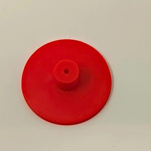 May include: A bright red, circular object with a central, cylindrical protrusion. The object appears to be made of a solid material, possibly plastic or resin. The design is simple and functional, with a small hole in the center of the cylinder.