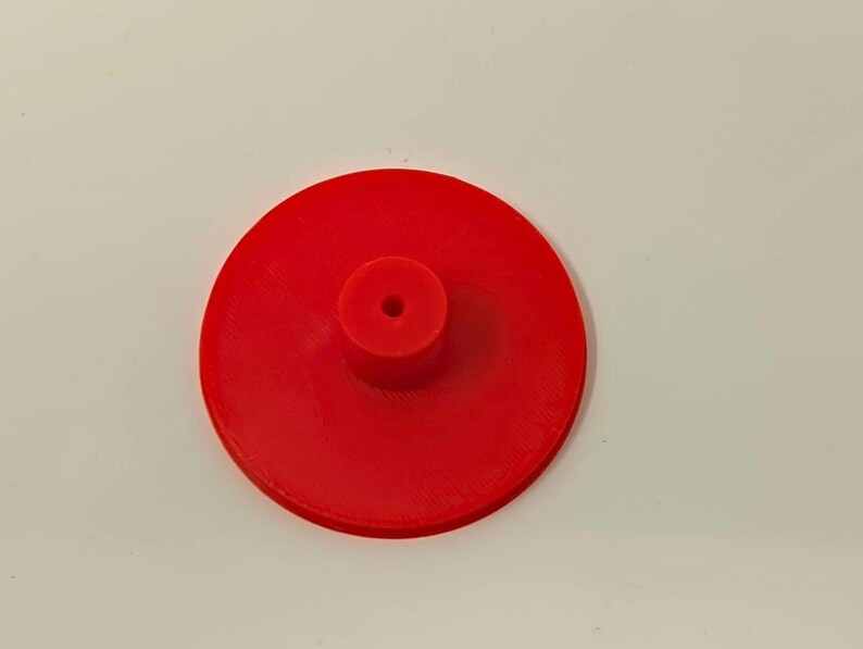 May include: A bright red, circular object with a central, cylindrical protrusion. The object appears to be made of a solid material, possibly plastic or resin. The design is simple and functional, with a small hole in the center of the cylinder.
