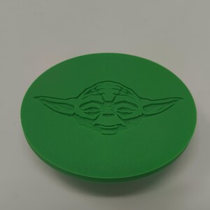 May include: A green, circular object featuring a detailed embossed image of Yoda's face from Star Wars. The design is centered on the disc, showcasing the character's distinctive features. The object is a solid, single-color item.