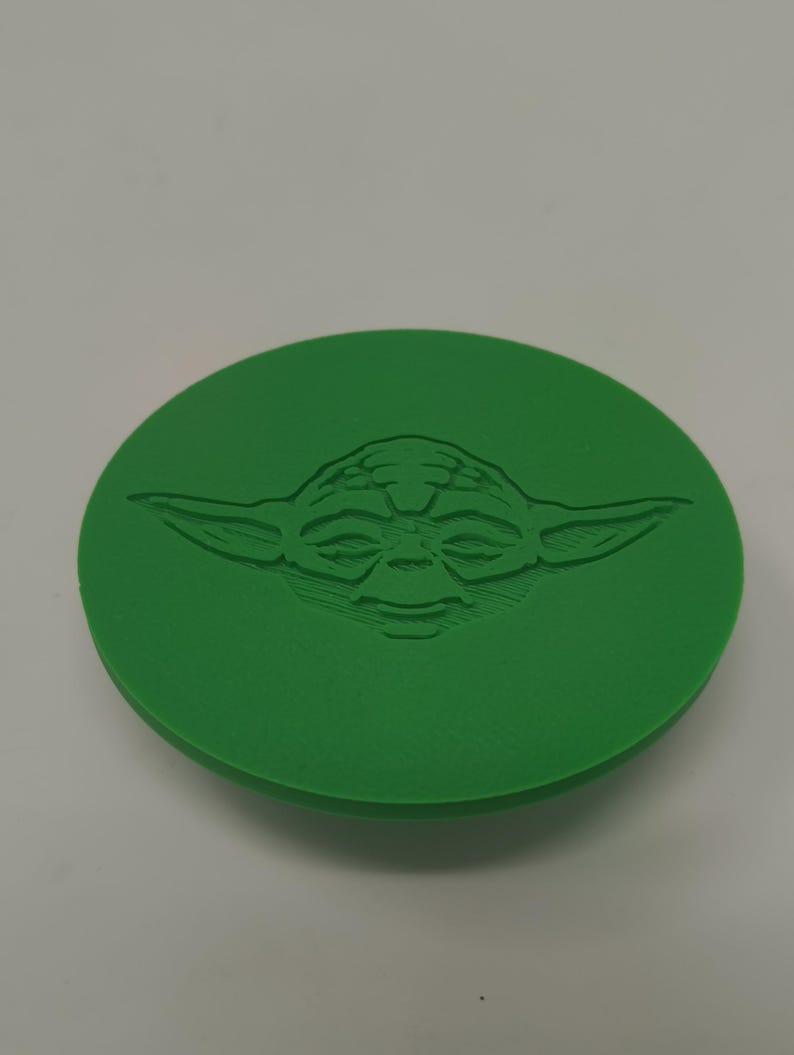 May include: A green, circular object featuring a detailed embossed image of Yoda's face from Star Wars. The design is centered on the disc, showcasing the character's distinctive features. The object is a solid, single-color item.