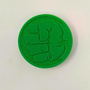 Puede incluir: Un botón redondo de plástico verde con una imagen en relieve de un puño verde.