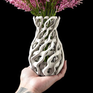 May include: A white, intricately designed vase with a unique, open-weave pattern. The vase holds a bouquet of pink flowers with green stems, set against a black background. The vase is held in a hand.