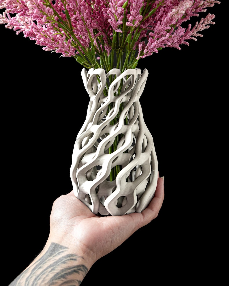 May include: A white, intricately designed vase with a unique, open-weave pattern. The vase holds a bouquet of pink flowers with green stems, set against a black background. The vase is held in a hand.