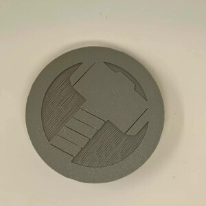 May include: A gray, circular emblem featuring a hammer design within a circle. The hammer is depicted with a textured surface, and the emblem has a raised border. The design is centered on a white background.