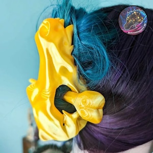 Könnte beinhalten: Eine leuchtend gelbe Plastik-Haarklammer in Form eines Drachenschädels. Die Klammer wird verwendet, um das blaue und violette Haar einer Person zu halten.