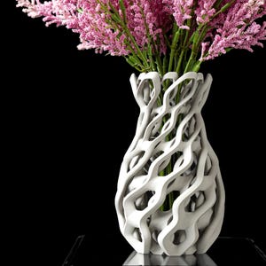 May include: A white, intricately designed vase holds a bouquet of pink flowers with green stems. The vase has a unique, open, woven pattern. The background is solid black, highlighting the vase and flowers.