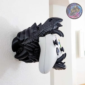 Puede incluir: Soporte de pared con cabeza de dragón negro que sostiene un mando de videojuegos blanco. La cabeza del dragón tiene escamas texturizadas y dientes afilados. El mando tiene botones y joysticks negros. Una pegatina con un logotipo es visible.