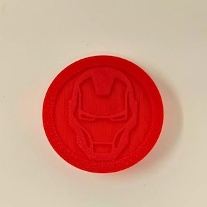 Pode incluir: Um emblema vermelho e circular com o design do capacete do Homem de Ferro. O emblema é um objeto sólido tridimensional com uma superfície texturizada. O design é um símbolo de super-herói reconhecível.