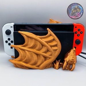 May include: A gold 3D printed dragon stand for a Nintendo Switch console. The dragon has wings and a tail, and the console sits in the wings. The console is black and red, and the stand is on a white surface.