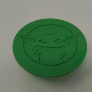 May include: A green, circular object featuring a raised relief of a character with large ears and wide eyes. The character is centered within a circular border. The object is a solid, vibrant green color.