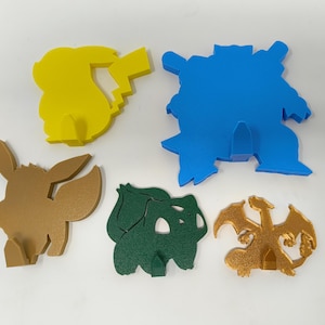 Könnte beinhalten: Fünf verschiedene Pokemon-Charakter-Wandhaken aus Kunststoff. Die Haken haben die Form von Pikachu, Eevee, Glurak, Schiggy und Bisasam. Die Haken sind alle in verschiedenen Farben, darunter Gelb, Braun, Gold, Blau und Grün.