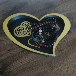 May include: A heart-shaped decorative tray with a black interior and gold trim. The tray contains several beaded necklaces in various colors. The tray has a decorative heart design on the inside.