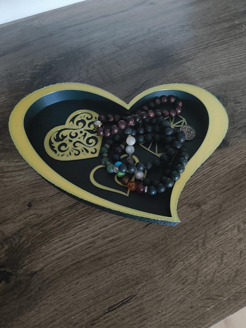 May include: A heart-shaped decorative tray with a black interior and gold trim. The tray contains several beaded necklaces in various colors. The tray has a decorative heart design on the inside.