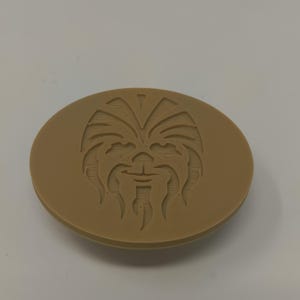 May include: A tan-colored, oval-shaped object with a detailed relief of a Wookiee face. The design features the character's distinctive facial features, including eyes, nose, and mouth. The object is likely a decorative item or a collectible.