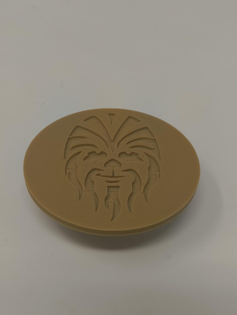 May include: A tan-colored, oval-shaped object with a detailed relief of a Wookiee face. The design features the character's distinctive facial features, including eyes, nose, and mouth. The object is likely a decorative item or a collectible.