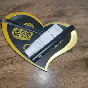 May include: A heart-shaped decorative tray with a black center and gold trim, holding makeup brushes, a tube of Laura Mercier moisturizer, and a black mascara tube. The tray has a gold filigree design in the center.