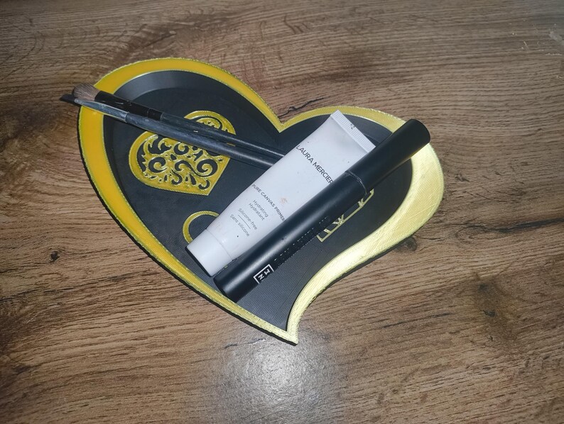 May include: A heart-shaped decorative tray with a black center and gold trim, holding makeup brushes, a tube of Laura Mercier moisturizer, and a black mascara tube. The tray has a gold filigree design in the center.