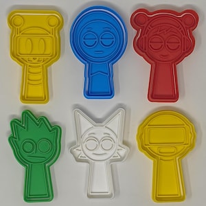 May include: Six colourful plastic cookie cutters in yellow, blue, red, green, white, and yellow. Each cutter features a cartoon character design with distinct facial features and shapes. The cutters are designed for making biscuits or other treats.