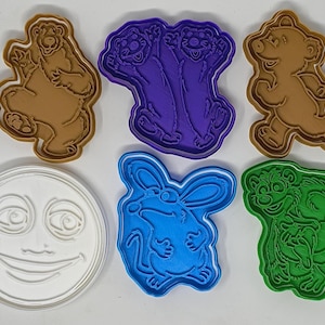 Puede incluir: Una colección de cortadores de galletas de plástico de colores en varias formas. Las formas incluyen una luna sonriente, un oso, un conejo y un personaje de dibujos animados. Los cortadores son en tonos marrón, morado, azul, verde y blanco.