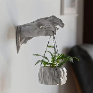 May include: A grey, wall-mounted hand sculpture holds a small, textured, grey planter filled with green ferns. The planter hangs from a chain. The sculpture is mounted on a white wall.