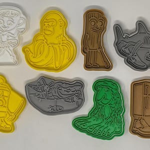 Könnte beinhalten: Eine Sammlung farbenfroher Ausstecher aus Kunststoff in verschiedenen Formen, darunter eine Ballerina, ein Gorilla, ein Tiki, ein Hai, ein Ninja, eine Landschaft, ein Troll und ein Kamel. Die Ausstecher sind in Weiß, Gelb, Braun, Grau und Grün gehalten.