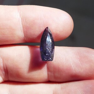 Puede incluir: Una pequeña gema facetada de color azul oscuro con una parte superior puntiaguda. La gema se sostiene en una mano, con el fondo desenfocado. La gema parece tener forma de bala.