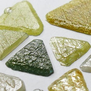 May include: A collection of twelve small, flat, triangular-shaped pieces in various shades of yellow, olive green, and metallic silver. Each piece features a textured, slightly irregular surface with distinct geometric edges and a glossy finish.