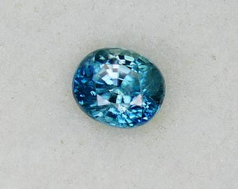 May include: An oval, blue gemstone sits in a white, square jewellery box. The gemstone displays a mix of light and dark blue shades, with a sparkling, textured surface. The box is set against a light brown wooden surface.
