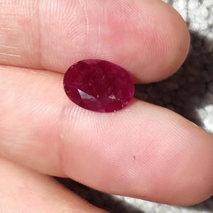 Può includere: Una gemma di rubino sfaccettata a forma ovale. Il rubino è di un colore rosso intenso e ricco e sembra lucido. La gemma è tenuta tra due dita su uno sfondo grigio chiaro strutturato.
