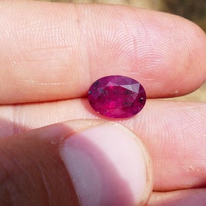Può includere: Una gemma di rubino rosso scuro a taglio ovale. La gemma sfaccettata è tenuta tra le dita, mostrando la sua superficie lucida e il suo colore vibrante. Le sfaccettature del rubino catturano la luce, mostrando la sua brillantezza.