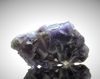 May include: A close-up of a fluorite crystal cluster. The crystal exhibits a complex, geometric structure with a mix of purple, gray, and white hues. The light catches the facets, creating a sparkling effect. The overall appearance is of a natural, geological specimen.