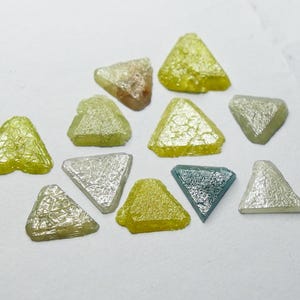 Pode incluir: Uma coleção de pedras preciosas triangulares em várias cores, incluindo amarelo, prata e verde-azulado. As pedras têm uma superfície texturizada e estão dispostas sobre um fundo branco. Provavelmente diamantes brutos.