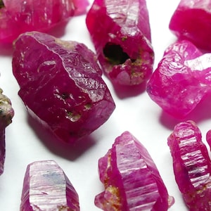 以下が含まれることがあります： さまざまな形とサイズの、未加工のルビーの結晶のコレクション。宝石は、自然な質感と不完全さが目に見える、深く鮮やかなピンクがかった赤色を示しています。一部の結晶はわずかに半透明に見えます。