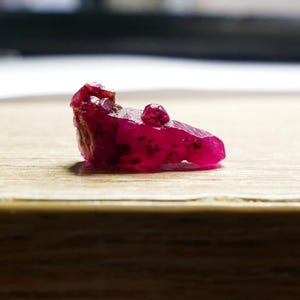 Op de afbeelding: Een ruwe, ongeslepen robijn edelsteen met een dieprode kleur. De robijn heeft een onregelmatige vorm met een ruw, getextureerd oppervlak en een kleine, afgeronde uitsteeksel. Het ligt op een lichtbruin houten oppervlak, met een vage achtergrond.