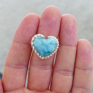 Könnte beinhalten: Ein herzförmiger Ring mit einem hellblauen Edelstein. Der Ring hat eine silberfarbene Metalleinfassung mit einem geflochtenen Seilmuster am Rand. Der Ring wird auf einer Hand gezeigt.