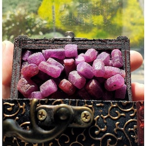 May include: A small, open wooden chest filled with raw, uncut ruby gemstones. The rubies are various shades of pink and red. The chest has a dark, ornate design on the exterior and is held in a person's hand.
