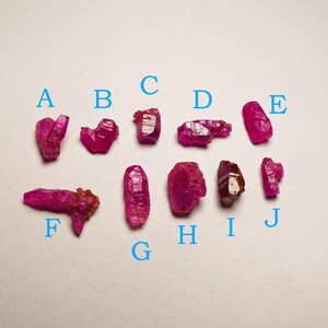 Puede incluir: Una colección de diez gemas de rubí en bruto, sin cortar, cada una con un vibrante tono rojo-rosado. Las gemas varían en tamaño y forma, con estructuras cristalinas visibles. Letras A a J en azul.