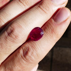 May include: A faceted, oval-shaped ruby gemstone. The ruby is a deep, saturated red color, with visible facets that catch the light. The gemstone is held in a hand, showcasing its size and brilliance.