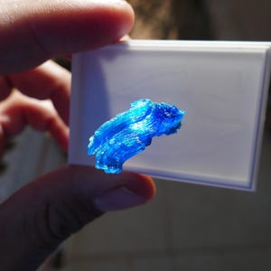 May include: A vibrant blue, crystalline structure is held against a white rectangular surface. The object has a textured appearance, with visible layers and a bright, almost glowing quality. The background is blurred, suggesting a close-up shot.
