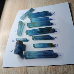 May include: A collection of eleven raw, elongated blue aquamarine crystals arranged on a white sheet of paper. Each crystal is labeled with a handwritten blue letter from A to K, showcasing varying shades of translucent blue with brown inclusions.